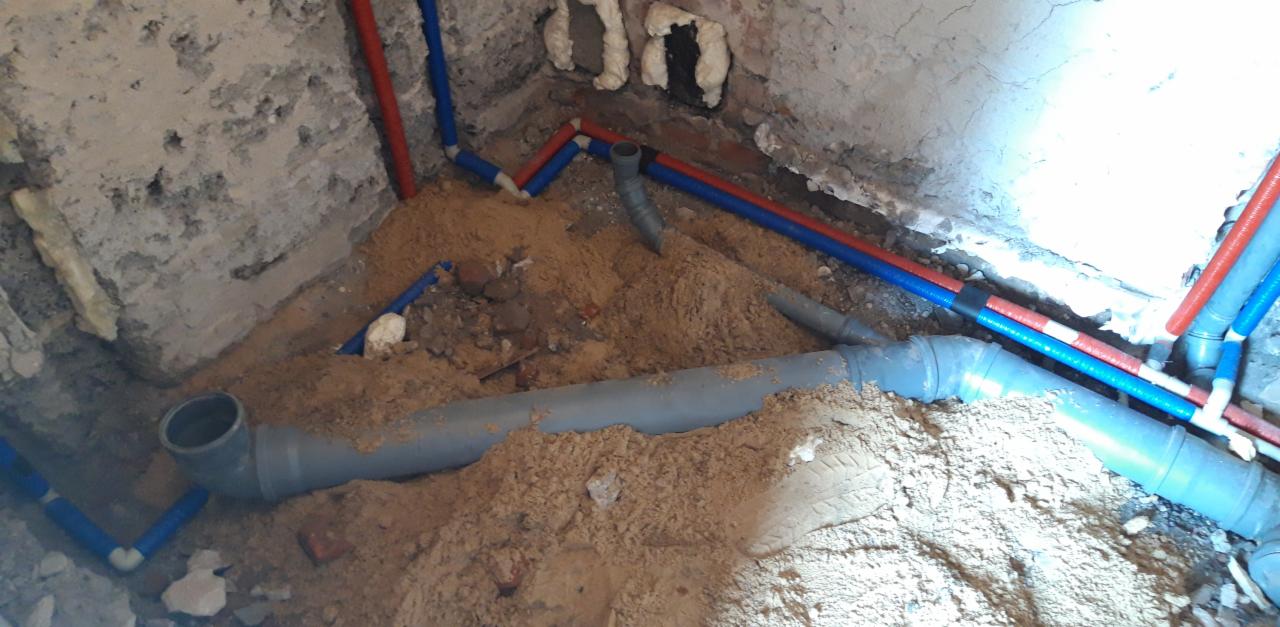 Instalacja wodno-kanalizacyjna w piasku, widoczne rury PCV szare i rury PEX czerwono-niebieskie w surowym wnętrzu budynku. Fragment instalacji w trakcie montażu.