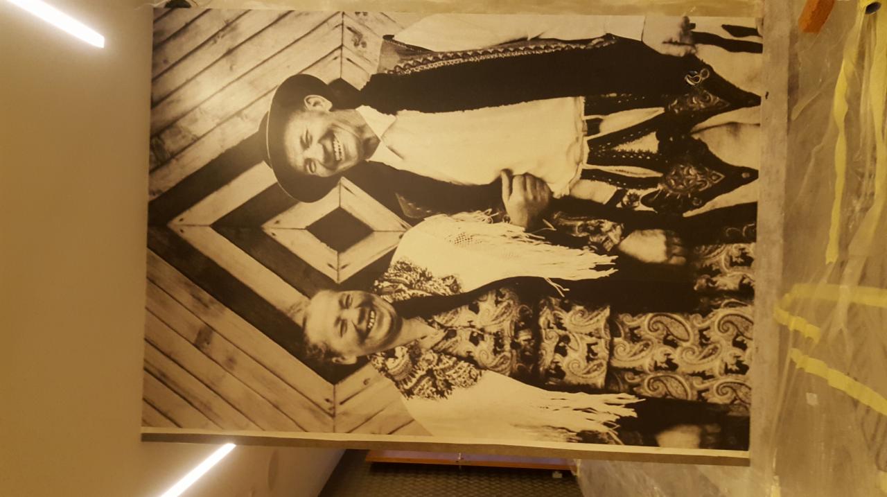 Duże, czarno-białe zdjęcie uśmiechniętej pary w strojach góralskich, naklejone na ścianę z drewnianą boazerią w romby, zabezpieczoną folią malarską.