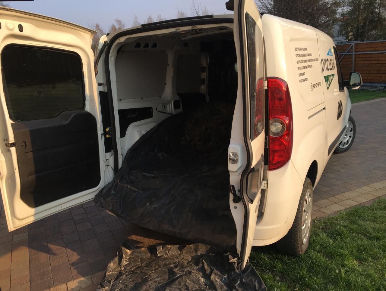 Otwarty bagażnik białego samochodu dostawczego wypełniony workiem z czarną folią zawierającym resztki roślinne, zaparkowanego na trawniku przed posesją.