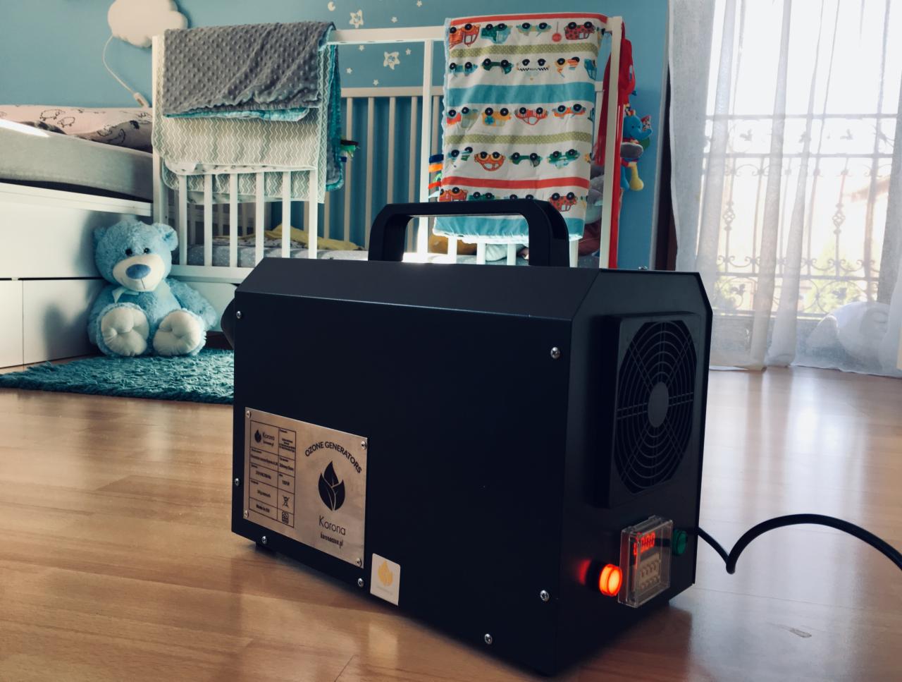 Czarny generator ozonu marki Korona na drewnianej podłodze w pokoju dziecięcym, z widocznym łóżeczkiem, pluszowym misiem i zasłonami w tle.
