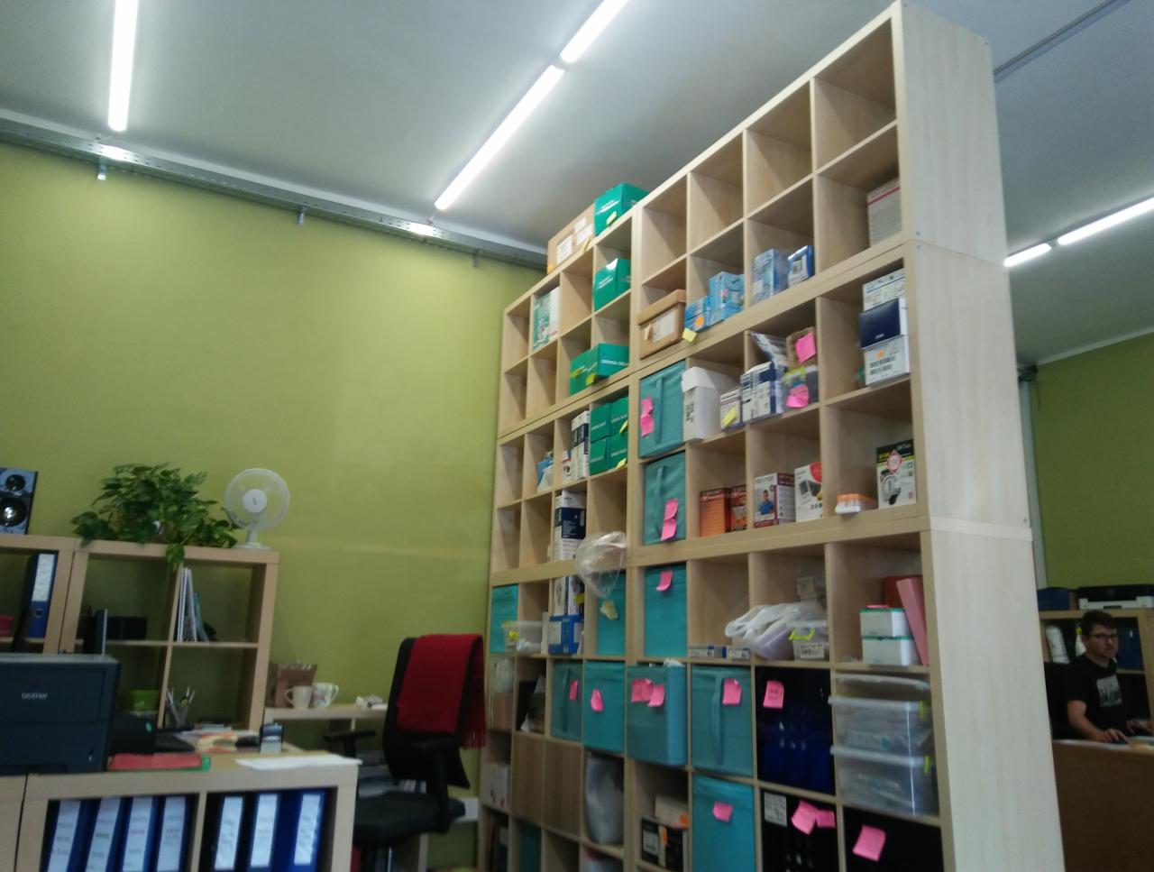 Magazyn z wysokim regałem wypełnionym pudełkami, pojemnikami i artykułami biurowymi, oznaczonymi różowymi karteczkami, widok z dołu w kierunku sufitu z lampami.