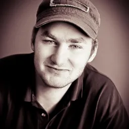 Portret mężczyzny w czapce z daszkiem, wykonany w sepii. Uśmiechnięta twarz, lekki zarost, ciemna koszula. Stonowane tło.