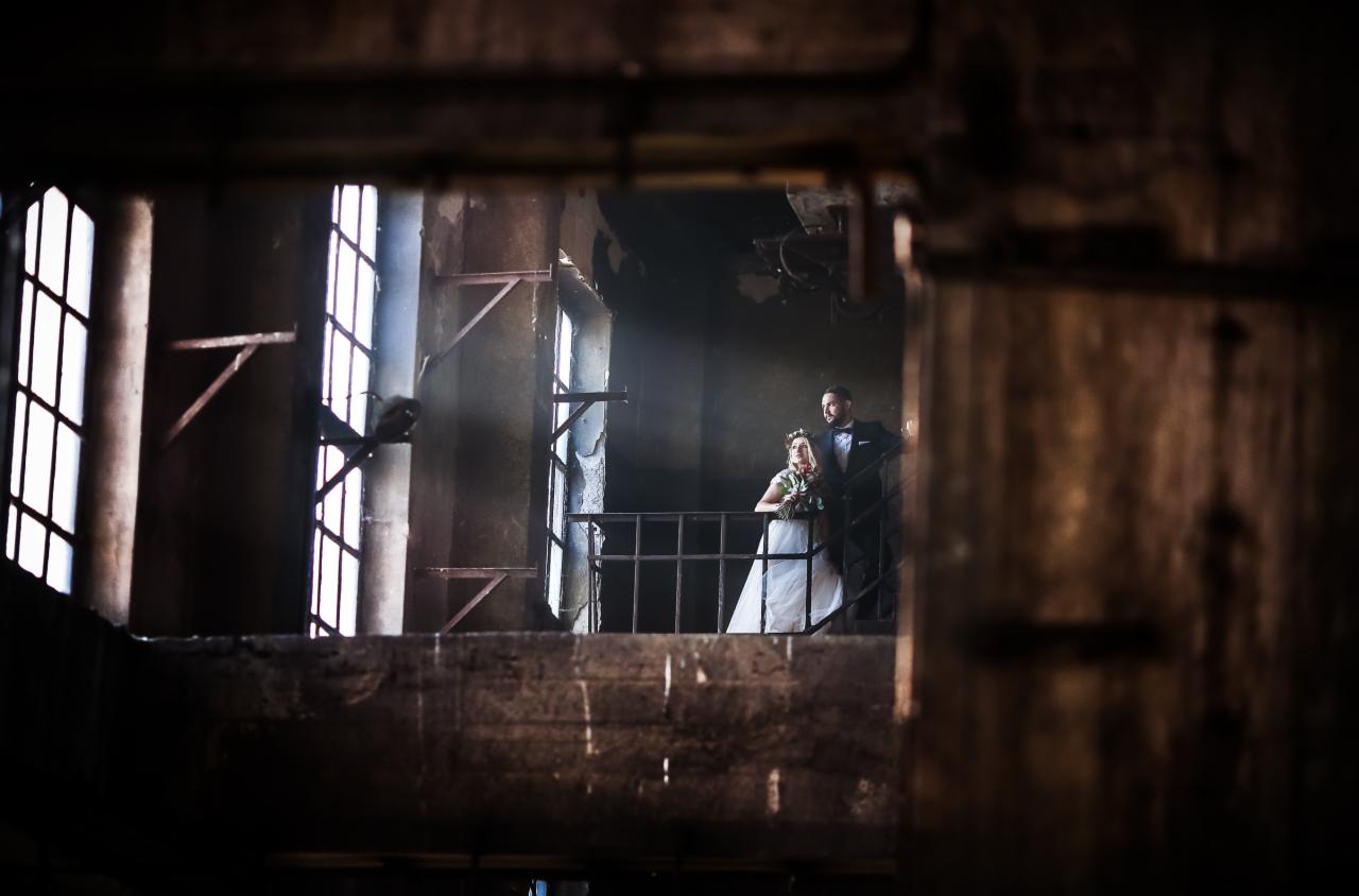 Para młoda na tle industrialnego wnętrza, widoczna przez ramę z drewnianych belek i metalowych elementów, światło wpadające przez okna tworzy dramatyczny efekt.