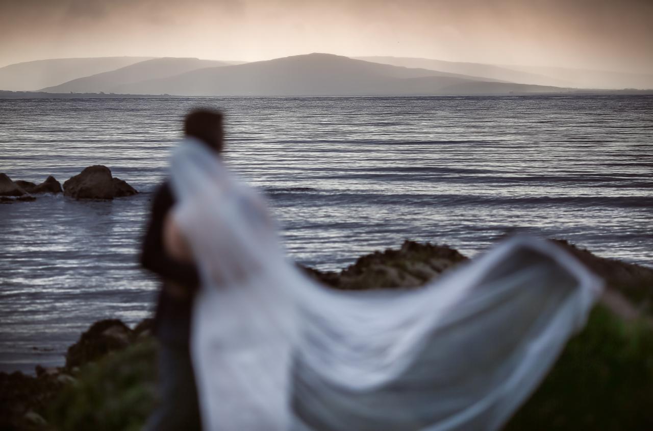 Para w objęciach na tle morza i wzgórz, z welonem na pierwszym planie, w stonowanej kolorystyce.