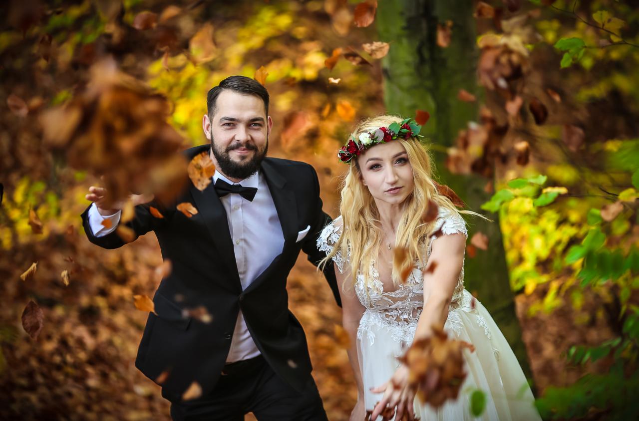Para młoda w lesie rzuca liśćmi, on w czarnym garniturze i muszce, ona w białej sukni z koronką i wiankiem z kwiatów.