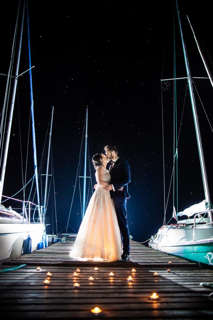 Pocałunek nowożeńców na molo oświetlonym świecami, w tle jachty i nocne niebo usiane gwiazdami.