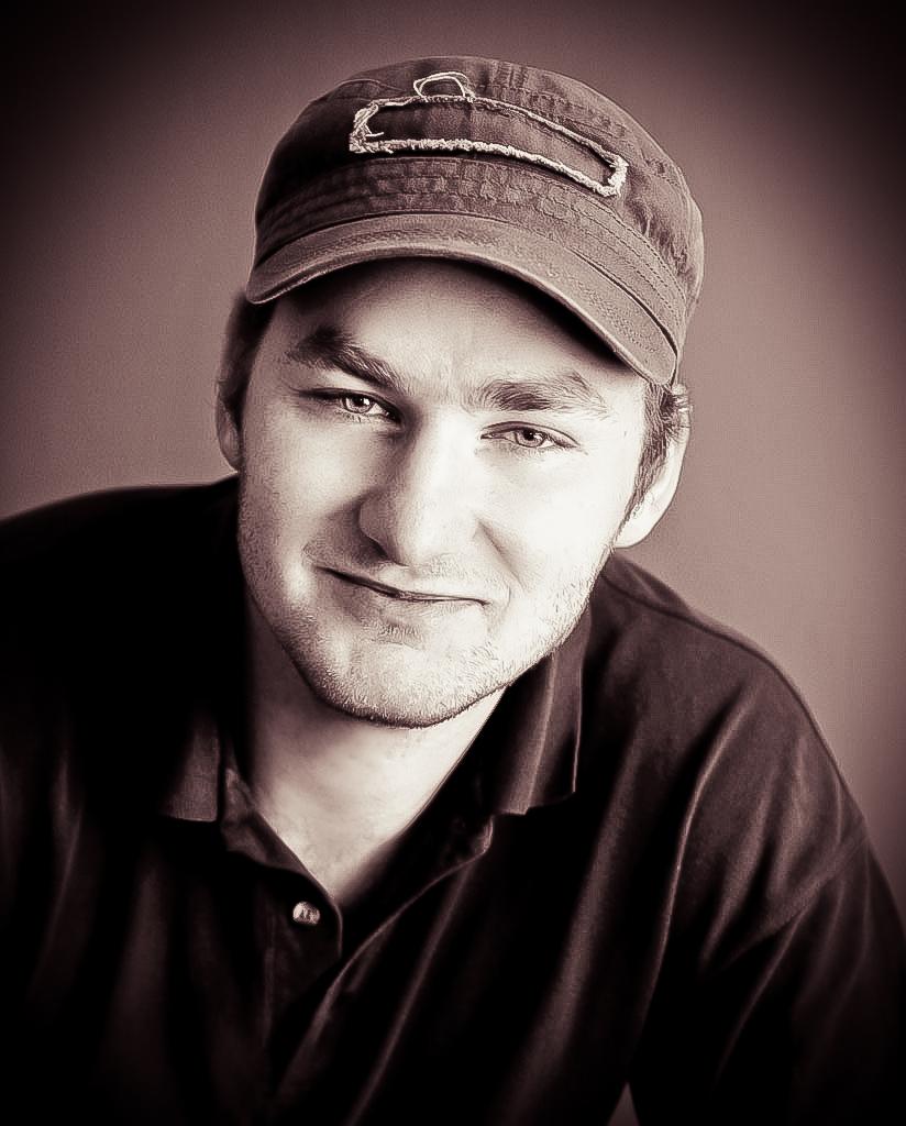 Portret mężczyzny w czapce z daszkiem, wykonany w sepii. Uśmiechnięta twarz, lekki zarost, ciemna koszula. Stonowane tło.