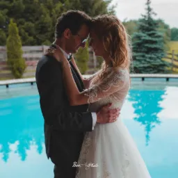 Para młoda w objęciach na tle basenu i zieleni, panna młoda w koronkowej sukni, pan młody w ciemnym garniturze i okularach.