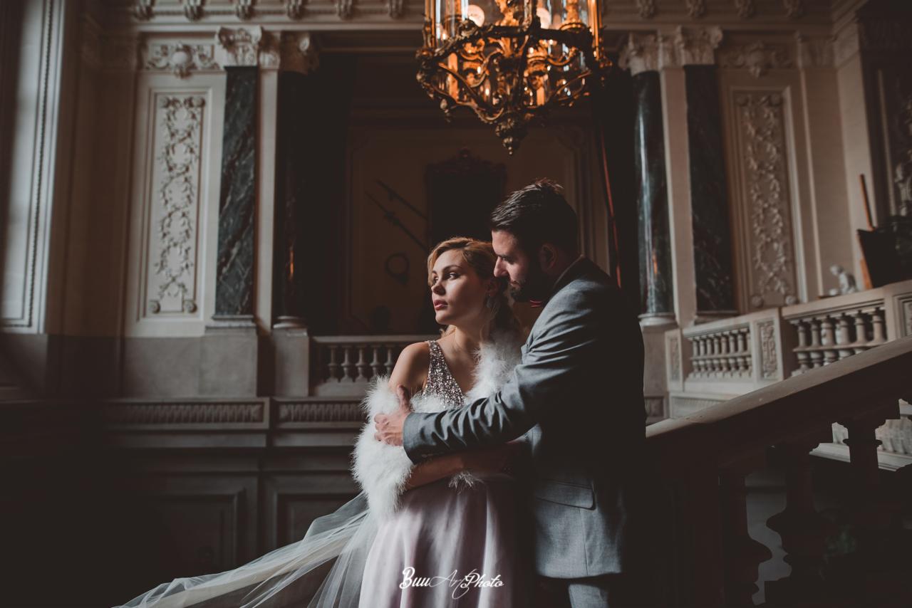 Elegancka para na schodach w zabytkowej kamienicy, kobieta w sukni z futrzanym szalem, mężczyzna w szarym garniturze, pod żyrandolem.