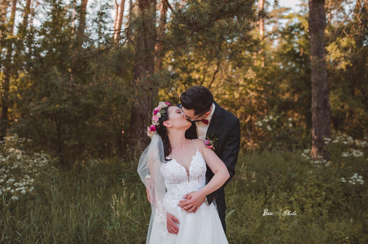 Para młoda całująca się na tle leśnej scenerii, panna młoda w koronie z kwiatów i sukni z koronkowym dekoltem, pan młody w ciemnym garniturze z bordową muchą.