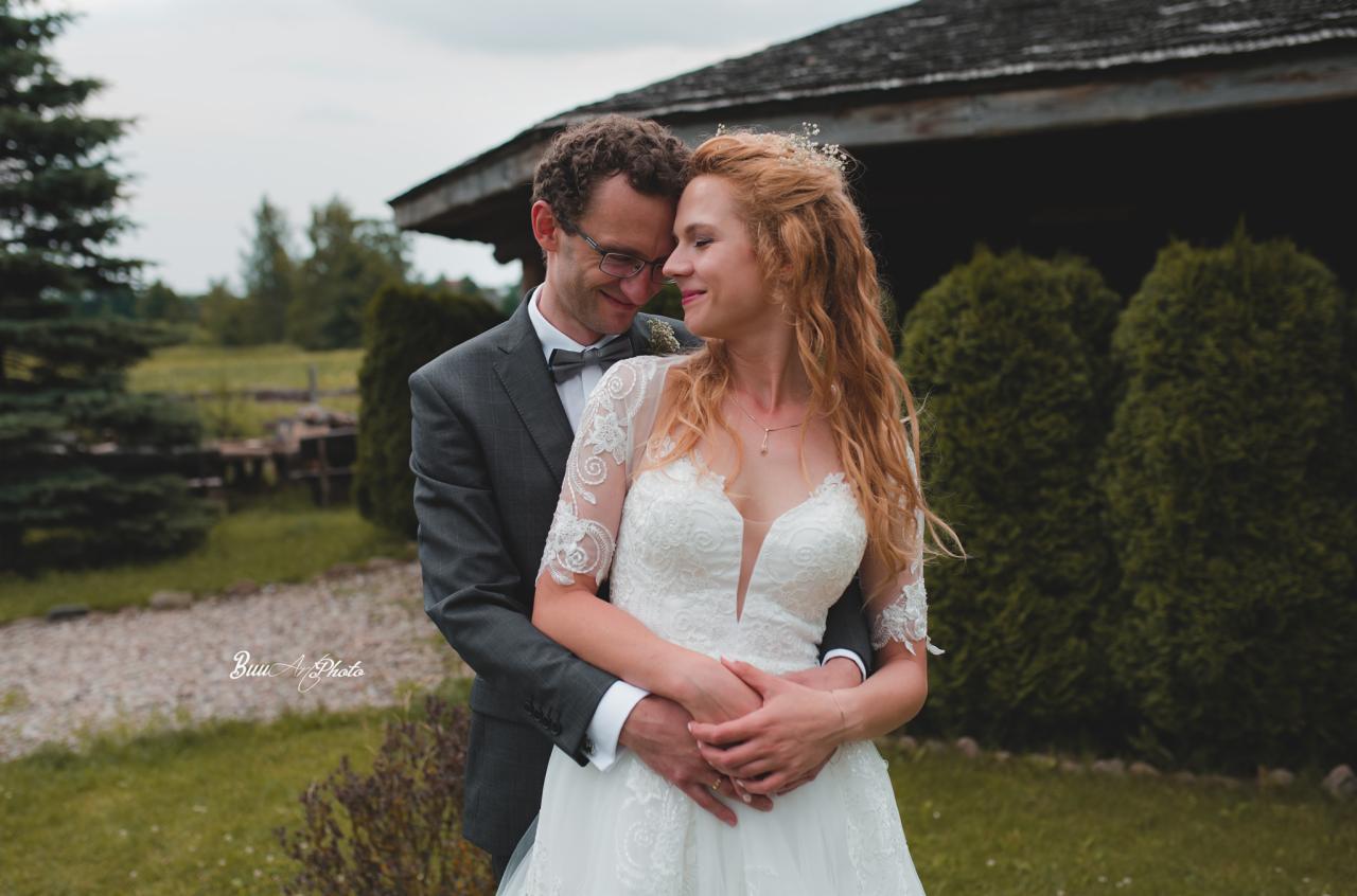 Para młoda w objęciach na tle zieleni i drewnianej konstrukcji; panna młoda w koronkowej sukni z dekoltem w serek, pan młody w szarym garniturze z muszką.