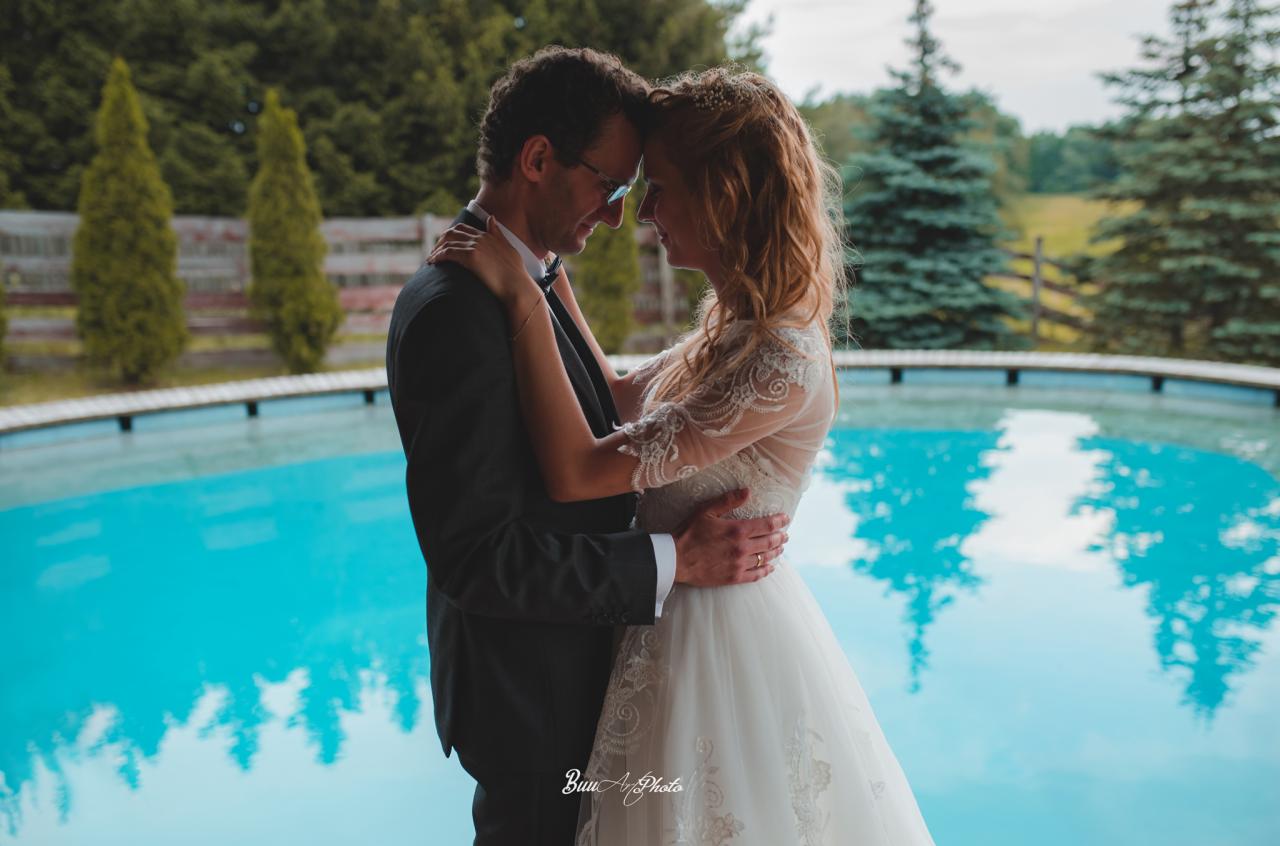 Para młoda w objęciach na tle basenu i zieleni, panna młoda w koronkowej sukni, pan młody w ciemnym garniturze i okularach.