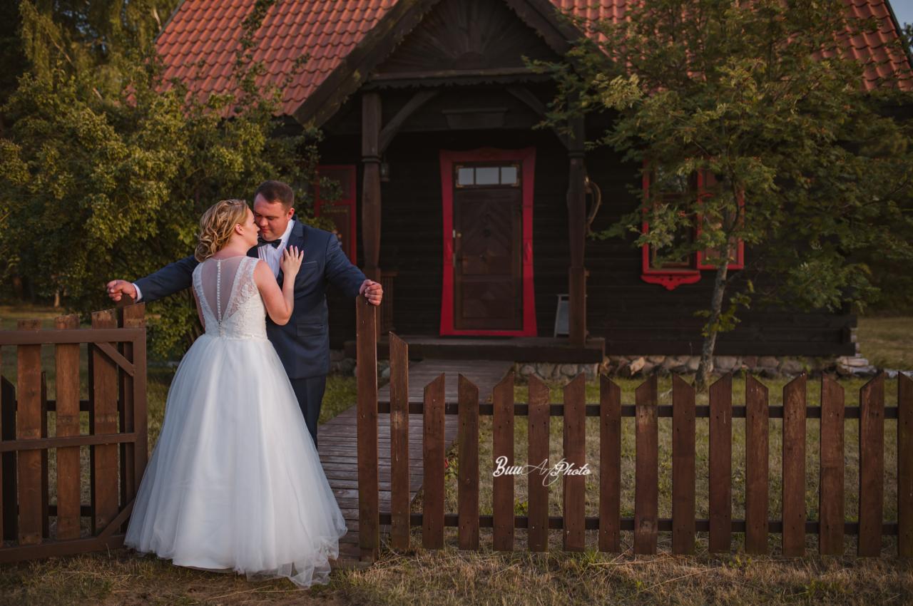 Para młoda w objęciach przy drewnianym płocie na tle ciemnego domu z czerwonymi ramami okiennymi i dachem z czerwonej dachówki.