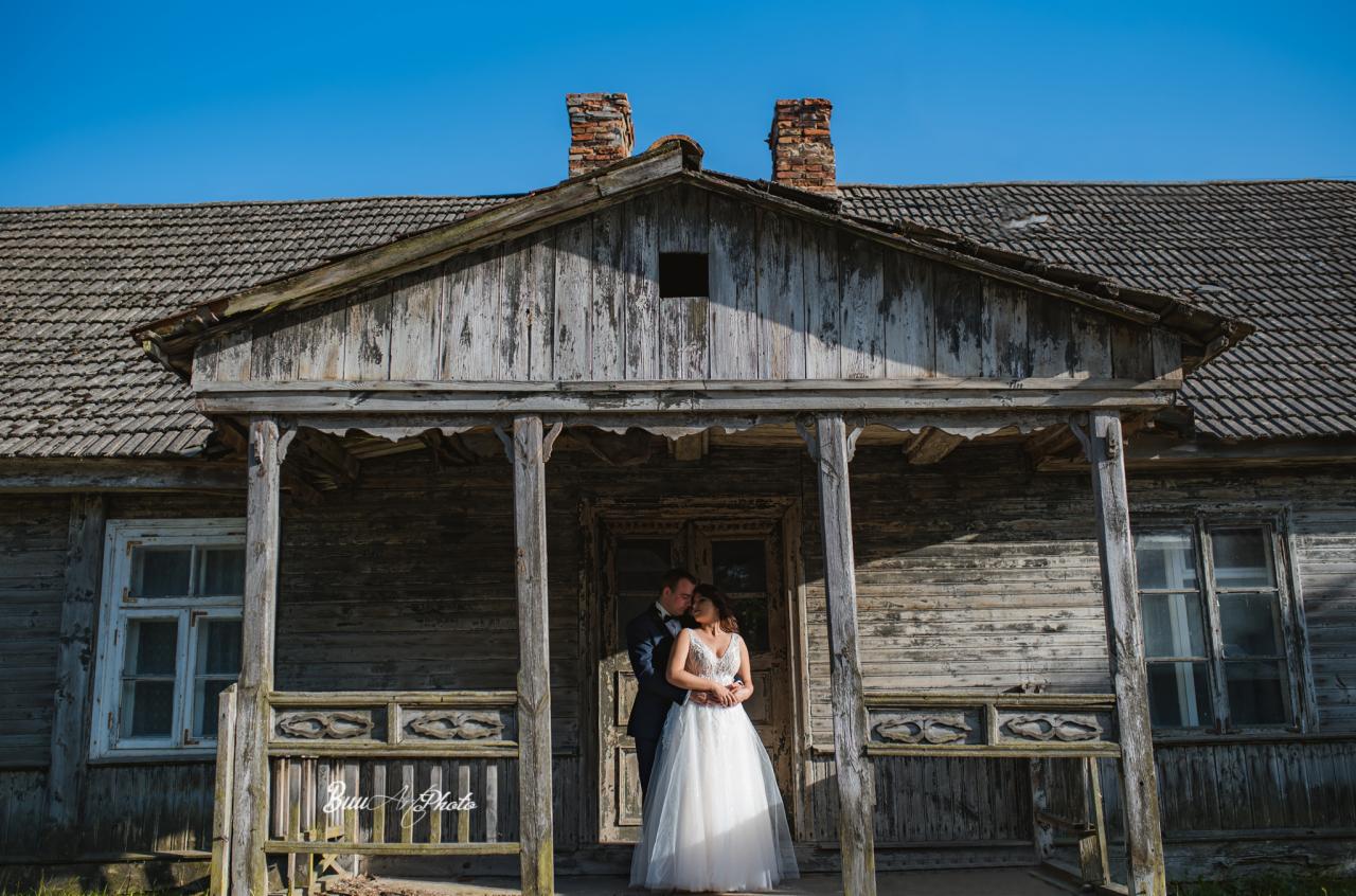 Para młoda w objęciach na tle starego, drewnianego domu z werandą i ceglanymi kominami, w słoneczny dzień.