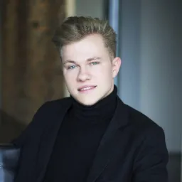 Młody mężczyzna o jasnych włosach i niebieskich oczach, ubrany w czarny golf i marynarkę, siedzi na czarnym fotelu. Portret w neutralnym oświetleniu.