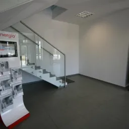 Wnętrze salonu sprzedaży z ekspozycją materiałów reklamowych, schodami ze szklaną balustradą i plakatem reklamowym w tle.