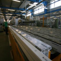 Elwiz S.A - Produkcja profili okiennych z tworzywa sztucznego w hali fabrycznej, widoczne długie rzędy profili Aluplast na taśmie produkcyjnej, pracownik w zielonej koszulce nadzorujący proces.