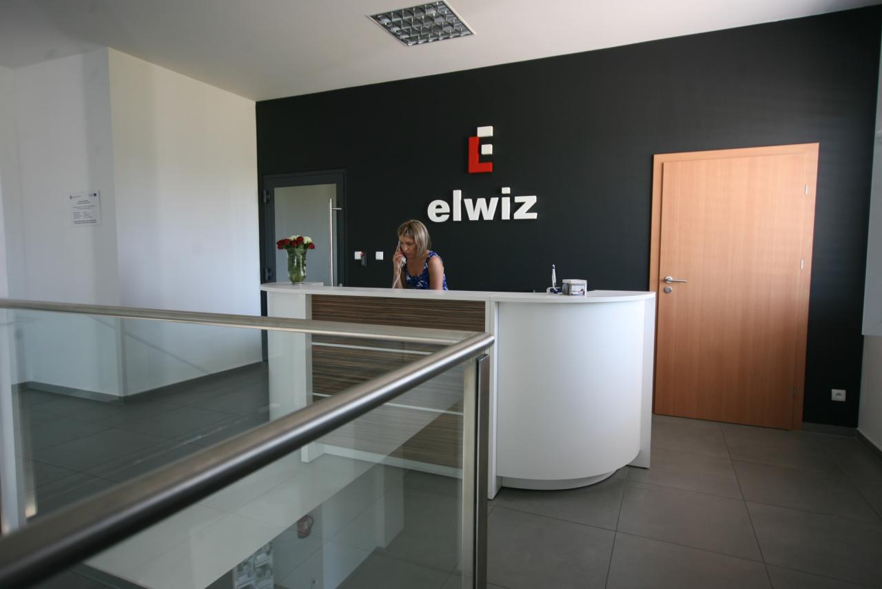 Recepcja firmy Elwiz z drewnianymi drzwiami po prawej, pracownica rozmawia przez telefon za białą ladą, widok z góry przez szklaną barierkę.