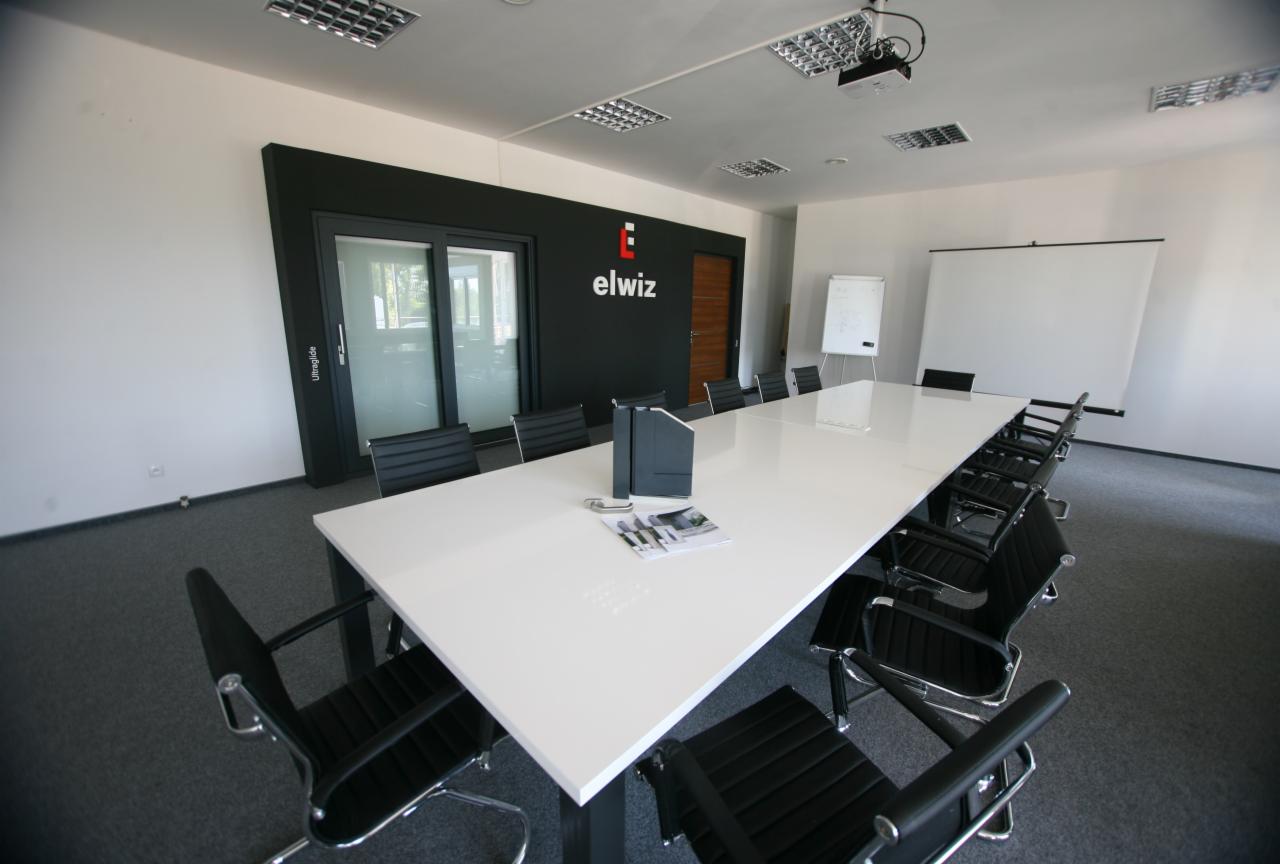 Sala konferencyjna z długim białym stołem, czarnymi krzesłami, tablicą suchościeralną i przesuwnymi drzwiami szklanymi z czarną ramą oraz logo firmy Elwiz na ścianie.