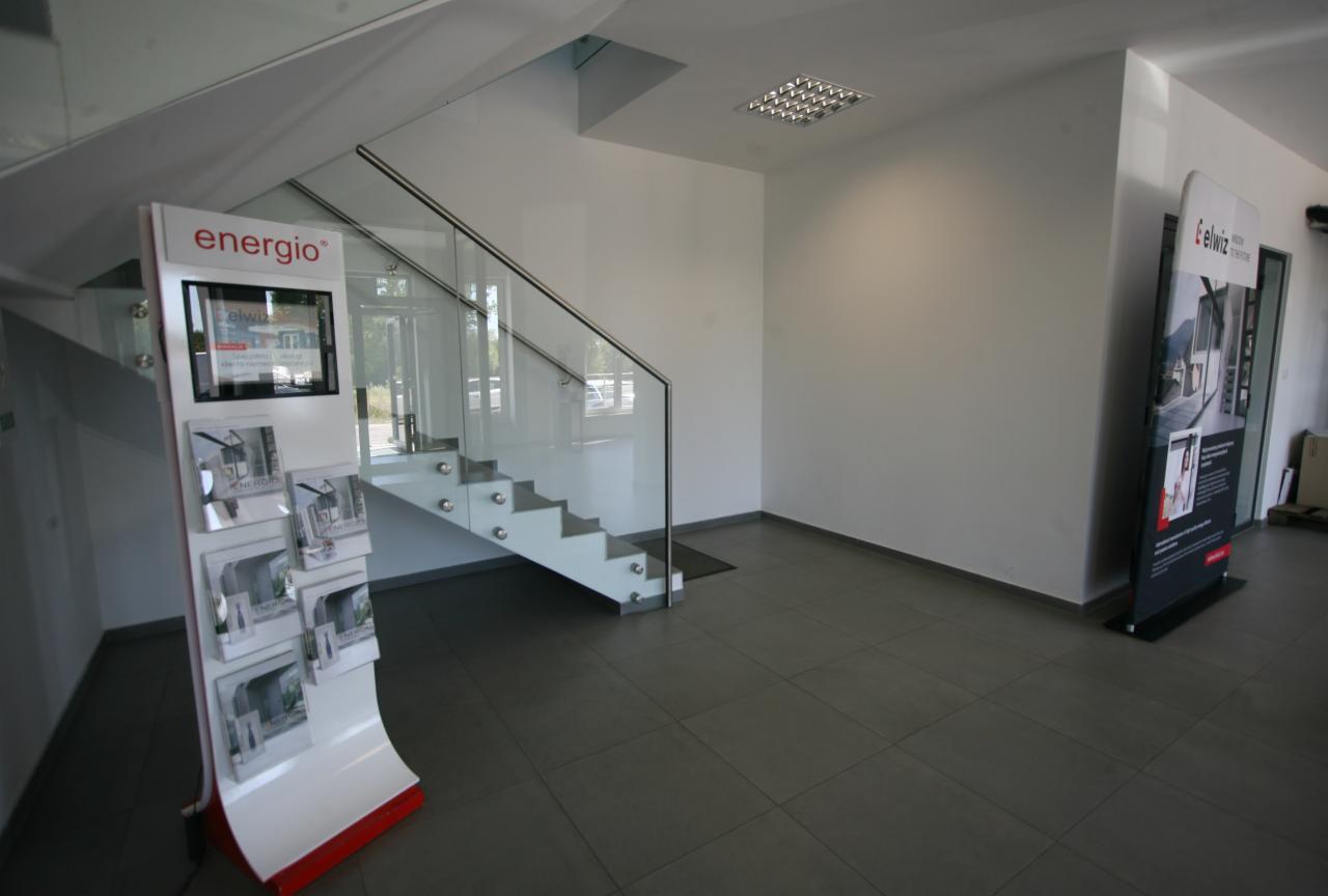 Wnętrze salonu sprzedaży z ekspozycją materiałów reklamowych, schodami ze szklaną balustradą i plakatem reklamowym w tle.