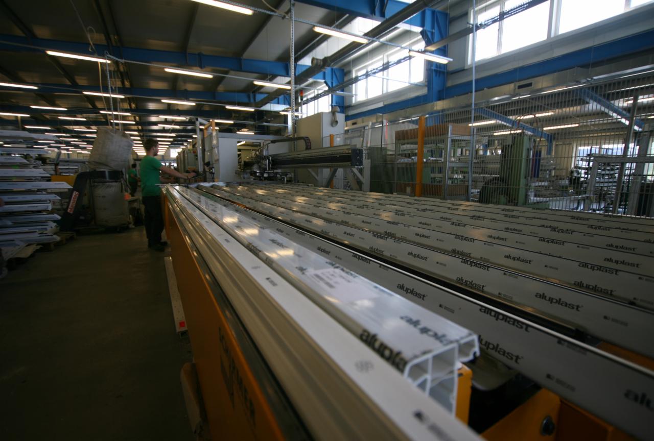 Produkcja profili okiennych z tworzywa sztucznego w hali fabrycznej, widoczne długie rzędy profili Aluplast na taśmie produkcyjnej, pracownik w zielonej koszulce nadzorujący proces.