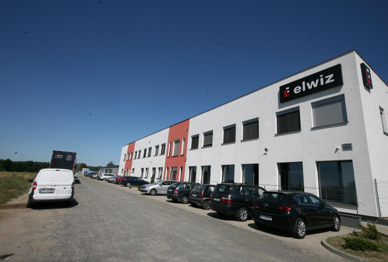 Budynek firmy z logo 'elwiz' na fasadzie, widoczne okna z żaluzjami zewnętrznymi i zaparkowane samochody.