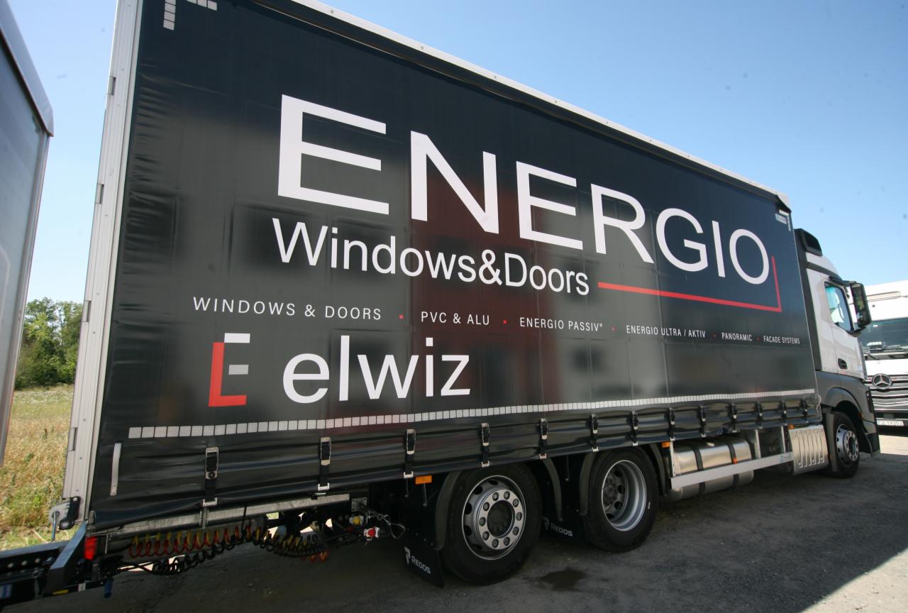 Ciężarówka z reklamą firmy produkującej okna i drzwi, z widocznym logo 'ENERGIO Windows&Doors' oraz 'elwiz' na czarnej plandece, zaparkowana na placu.