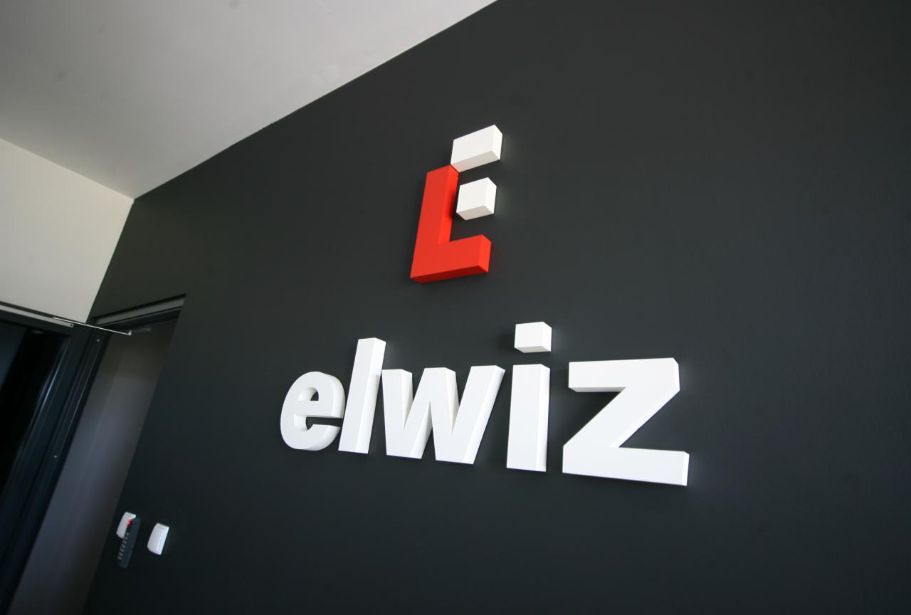 Logo firmy Elwiz, składające się z czerwono-białego symbolu graficznego oraz białego napisu, umieszczone na ciemnoszarej ścianie wewnątrz budynku, obok wejścia z widocznym fragmentem drzwi.