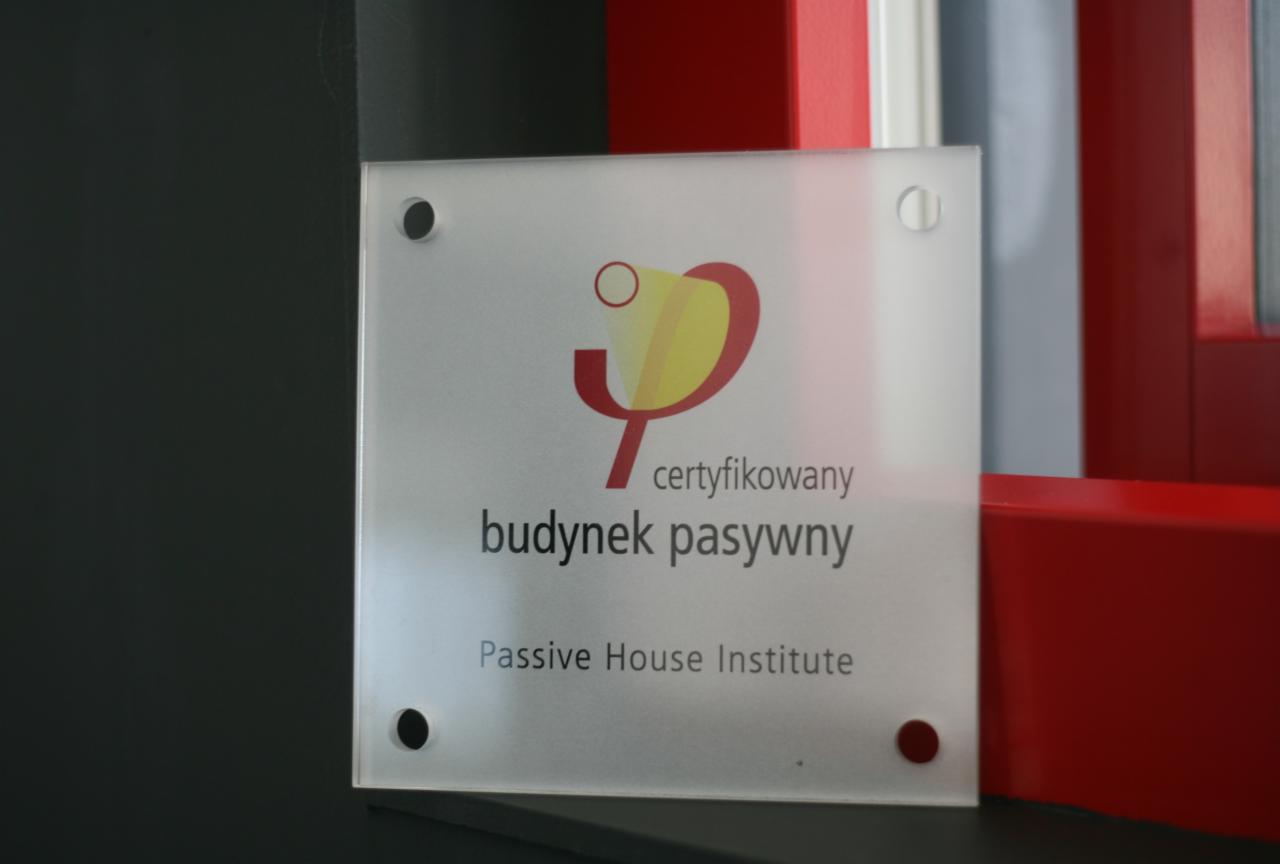 Tabliczka informacyjna 'certyfikowany budynek pasywny', umieszczona obok czerwonej ramy okiennej, z widocznym logo Passive House Institute.