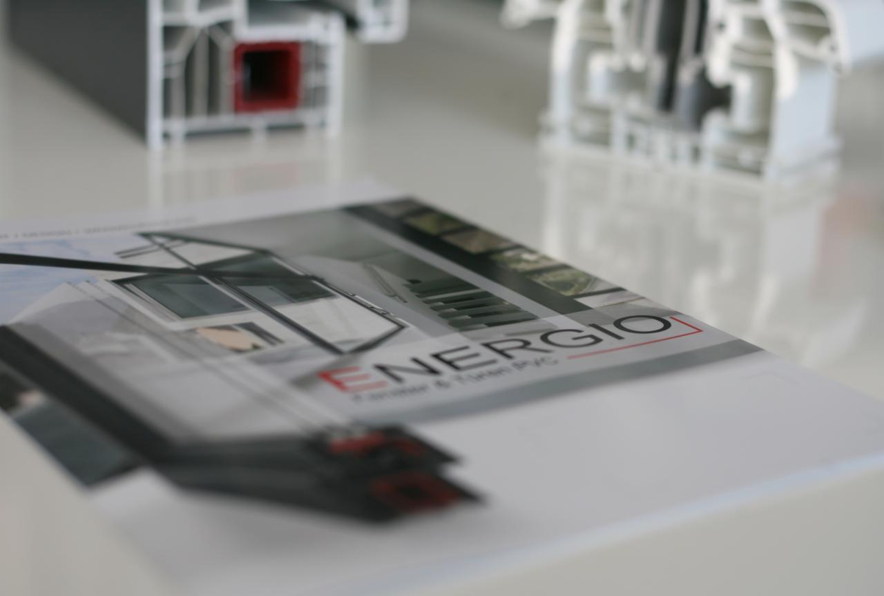 Przekrój profilu okiennego i katalog reklamowy z wizualizacją nowoczesnych okien w tle, leżące na białym, błyszczącym stole.