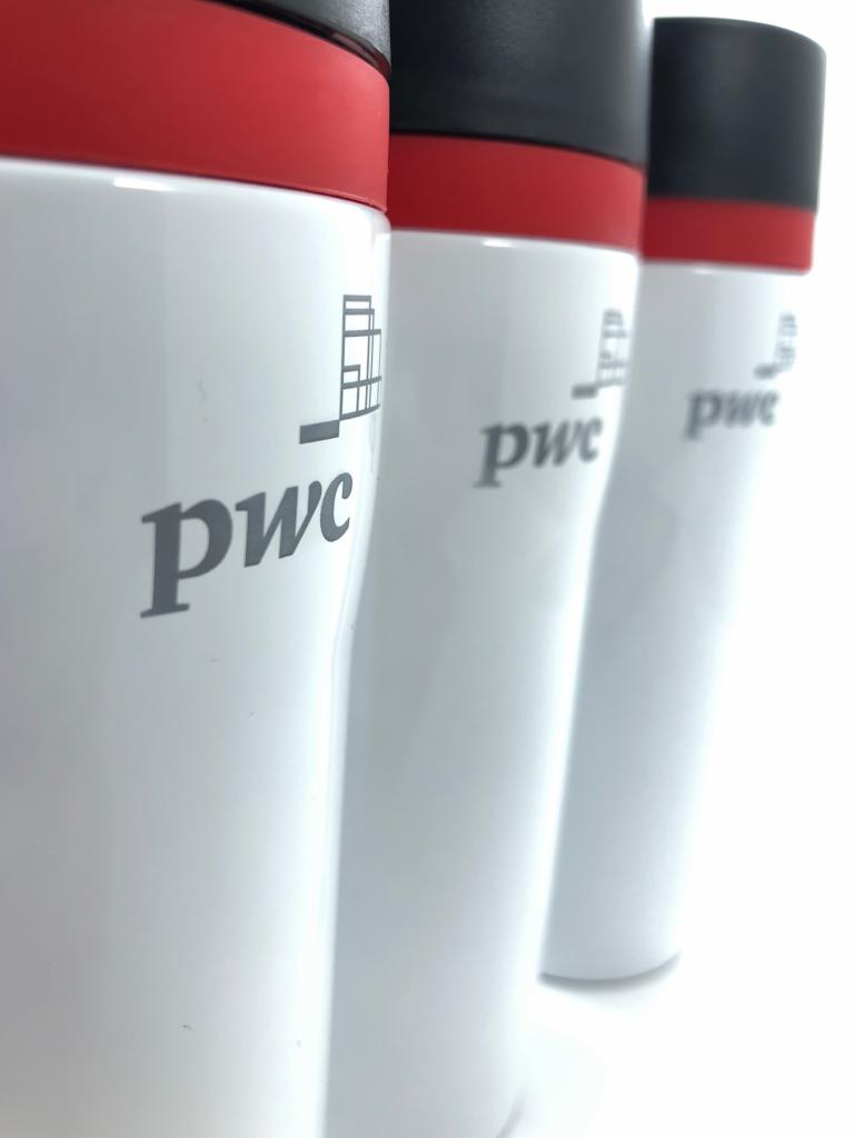 Trzy białe termokubki z logo PwC, z czerwonymi i czarnymi pokrywkami, ustawione w rzędzie na białym tle, widoczne z bliska.