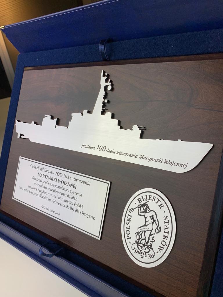 Pamiątkowa tablica z wyciętym laserowo profilem okrętu wojennego, dedykowana na 100-lecie Marynarki Wojennej, umieszczona w granatowym pudełku z tekstem okolicznościowym i logo Rejestru Statków.