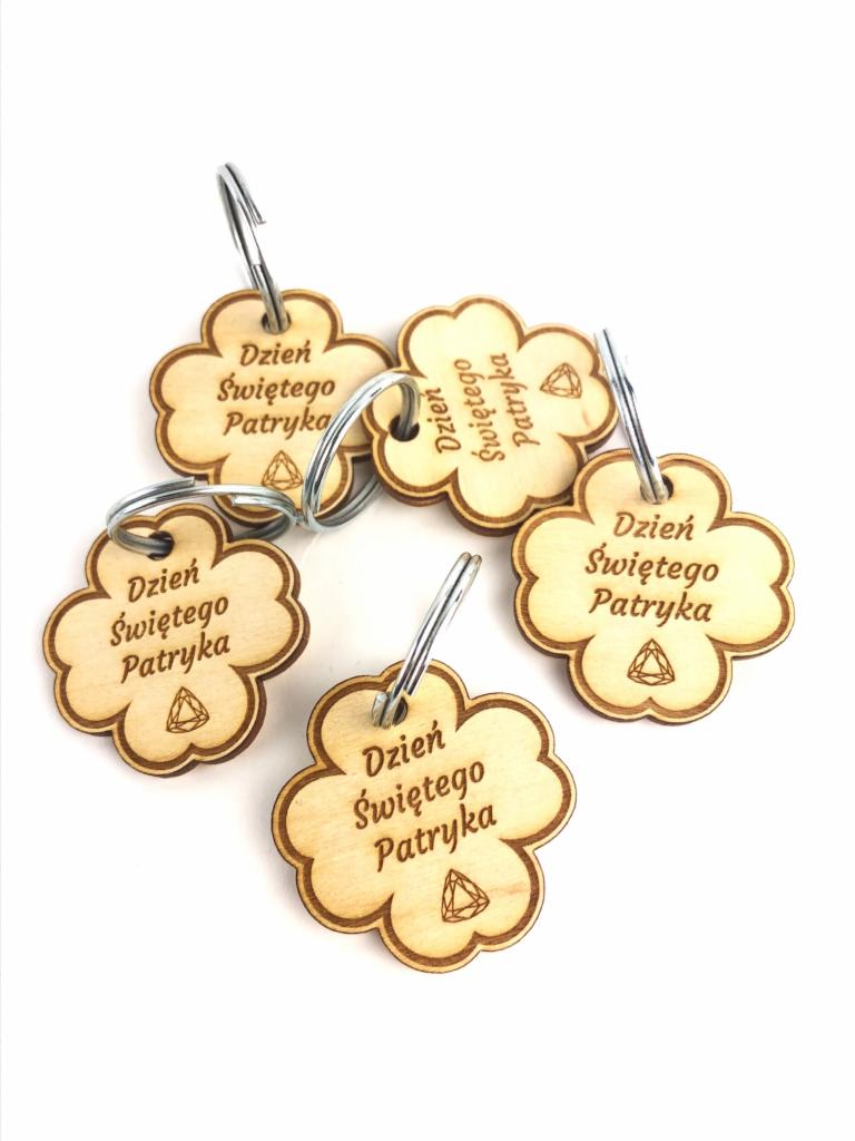 Zestaw pięciu drewnianych breloczków w kształcie koniczyny z grawerowanym napisem 'Dzień Świętego Patryka' i symbolem trójkąta, zawieszone na metalowych kółkach, na białym tle.