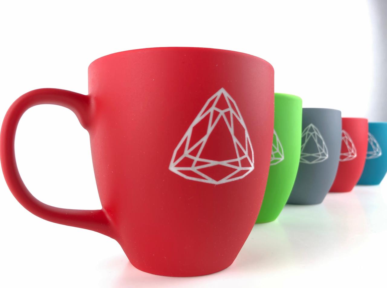 Rząd kolorowych kubków z logo, czerwony kubek na pierwszym planie, reszta kubków w odcieniach zieleni, szarości, czerwieni i błękitu, ustawione ukośnie na białym tle.