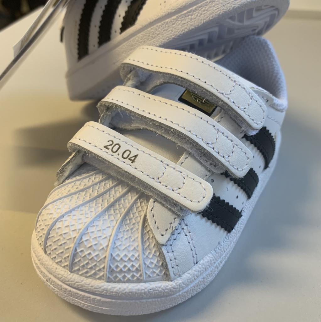Białe, dziecięce buty sportowe Adidas Superstar z trzema czarnymi paskami i zapięciem na rzepy, na jednym z pasków nadrukowana data '20.04'.