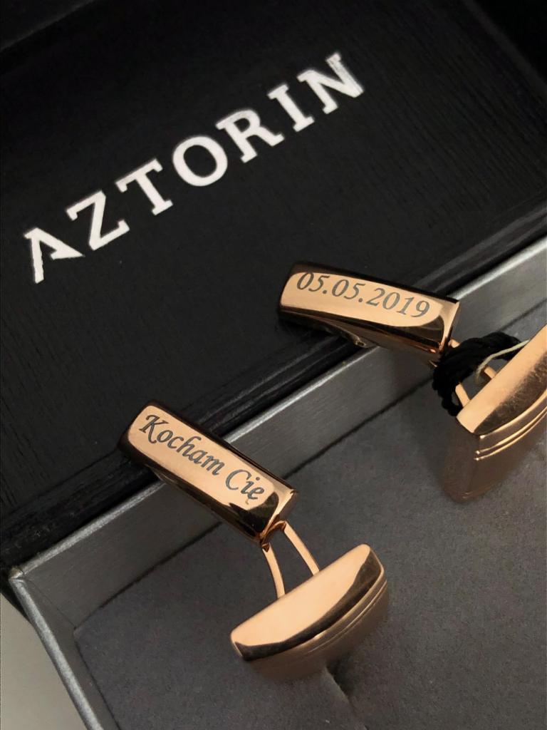 Różowe, pozłacane spinki do mankietów z grawerem 'Kocham Cię' i datą '05.05.2019' w czarnym pudełku prezentowym z logo 'AZTORIN'.
