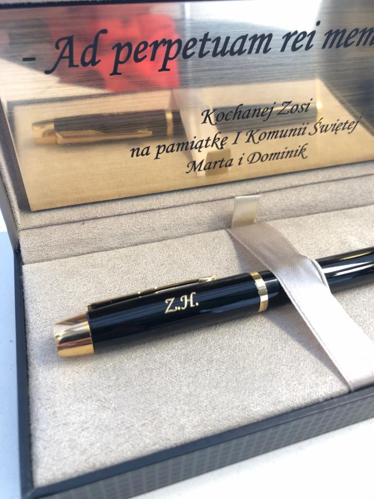 Elegancki, czarny długopis z grawerem inicjałów Z.H. w pudełku prezentowym z dedykacją 'Kochanej Zosi na pamiątkę I Komunii Świętej Marta i Dominik', całość przewiązana satynową wstążką.