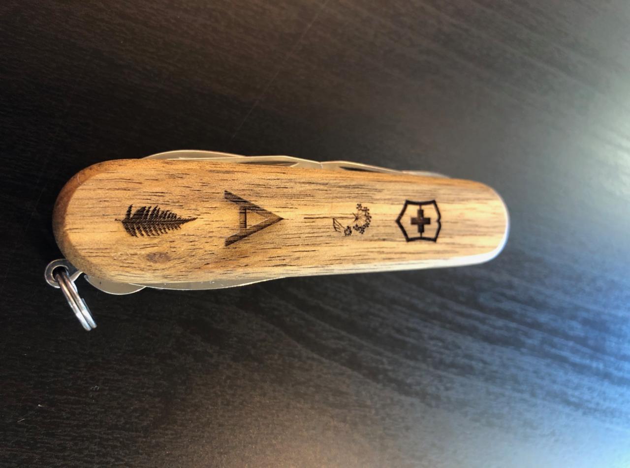 Drewniany scyzoryk z grawerowanymi symbolami: paproć, strzałka, dmuchawiec i krzyż, leżący na ciemnym blacie.