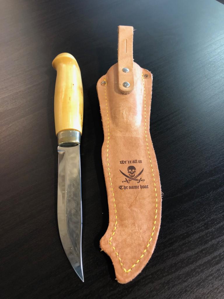 Nóż z jasną drewnianą rękojeścią leżący obok skórzanej pochwy z wygrawerowanym motywem pirackim i napisem 'We're all in The same boat'.