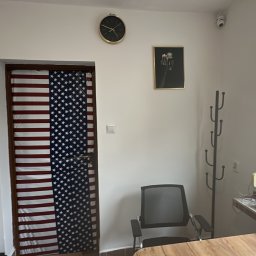 Anna Tajak - Sala lekcyjna z drzwiami ozdobionymi flagą USA, zegarem na ścianie i biurkiem przy oknie. Widoczny wieszak na ubrania i krzesło biurowe.