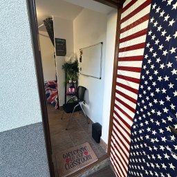 Anna Tajak - Wejście do klasy językowej z flagą USA na drzwiach i wycieraczką z napisem 'Welcome to our classroom'. W tle widoczny wystrój sali z flagą UK.
