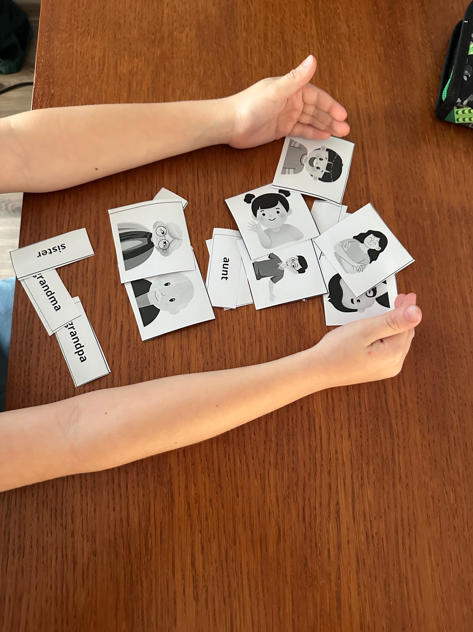 Dziecko układa karty z wizerunkami członków rodziny i podpisami (np. 'aunt', 'sister') na drewnianym stole. Widoczne dłonie dziecka i fragment plecaka.
