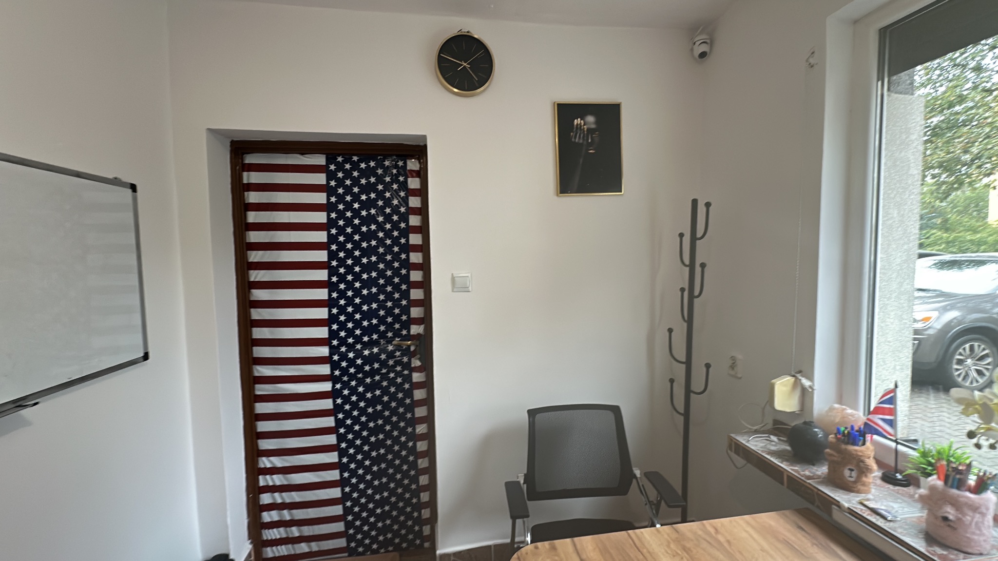 Sala lekcyjna z drzwiami ozdobionymi flagą USA, zegarem na ścianie i biurkiem przy oknie. Widoczny wieszak na ubrania i krzesło biurowe.