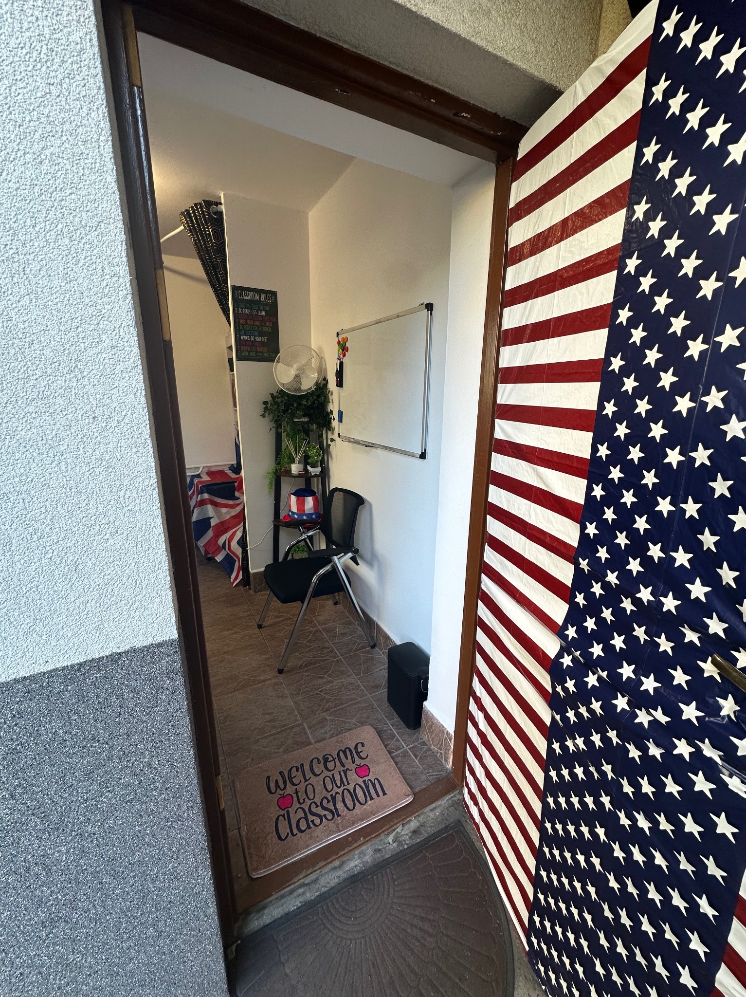 Wejście do klasy językowej z flagą USA na drzwiach i wycieraczką z napisem 'Welcome to our classroom'. W tle widoczny wystrój sali z flagą UK.