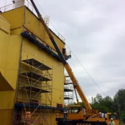 Żółty dźwig samochodowy z wysięgnikiem podnosi element na żółty budynek przemysłowy; widoczne rusztowania i komin w tle, pochmurne niebo.