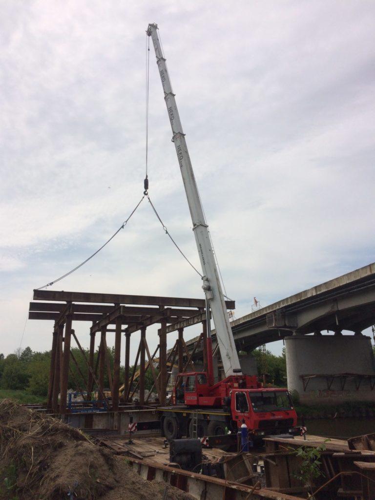 Czerwony dźwig teleskopowy marki NEKO podnosi stalową konstrukcję mostu na tle istniejącego betonowego wiaduktu i pochmurnego nieba.