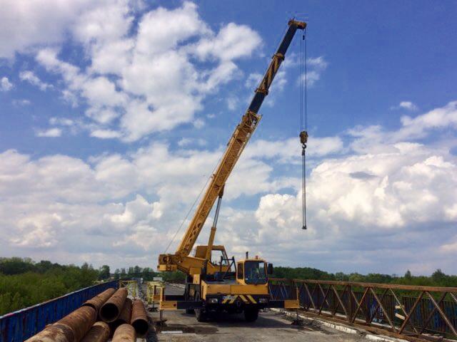Żółty dźwig teleskopowy na moście, podnosi rurę na tle błękitnego nieba z obłokami.