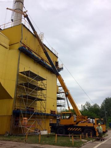 Żółty dźwig samochodowy z wysięgnikiem podnosi element na żółty budynek przemysłowy; widoczne rusztowania i komin w tle, pochmurne niebo.