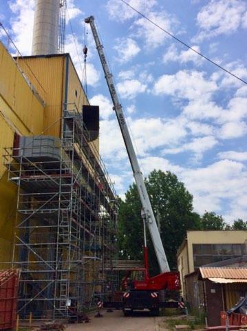 Żuraw samochodowy podnosi element konstrukcyjny na rusztowanie przy żółtym budynku przemysłowym z kominem, widoczne niebieskie niebo z chmurami.