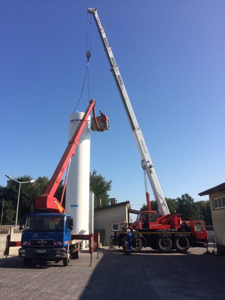 Dwa dźwigi, jeden z podnośnikiem koszowym, transportują biały, cylindryczny zbiornik na tle błękitnego nieba.